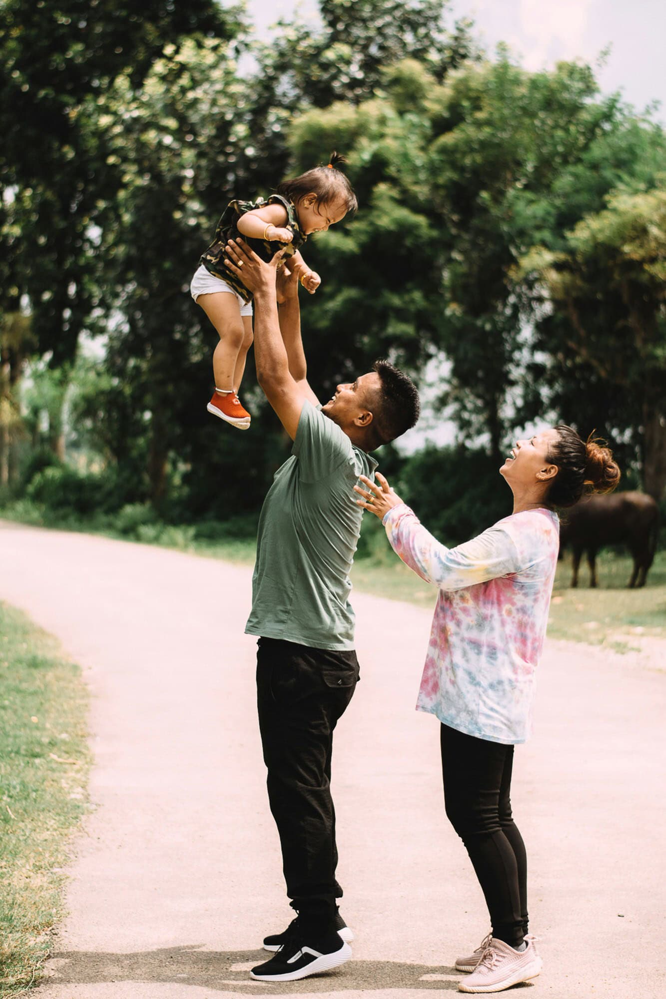 A man lifts a smiling toddler into the air while a woman stands next to them on a paved path surrounded by trees.