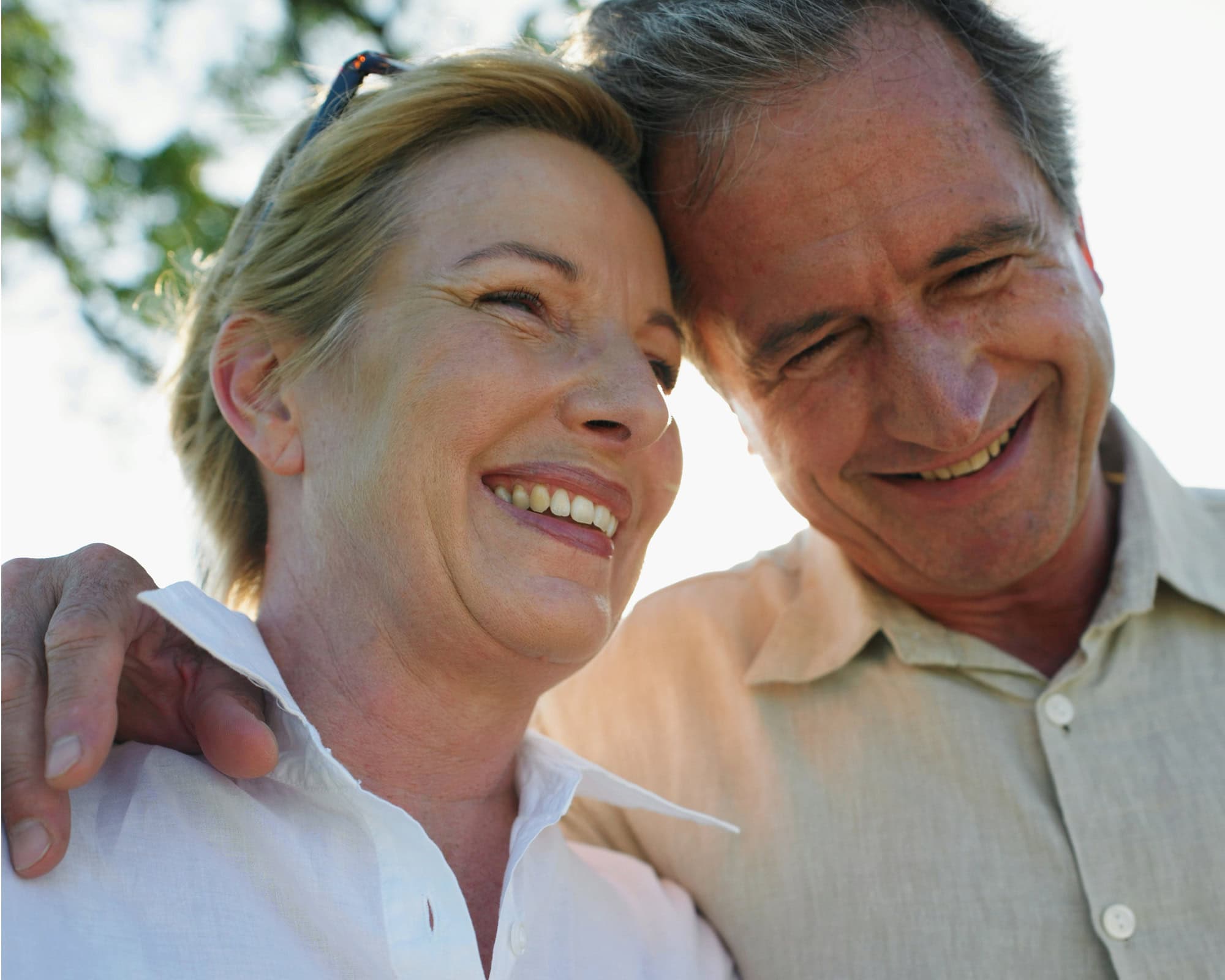 Two older adults, a woman and a man, stand close together outdoors, smiling and appearing happy. The man has his arm around the woman's shoulder.