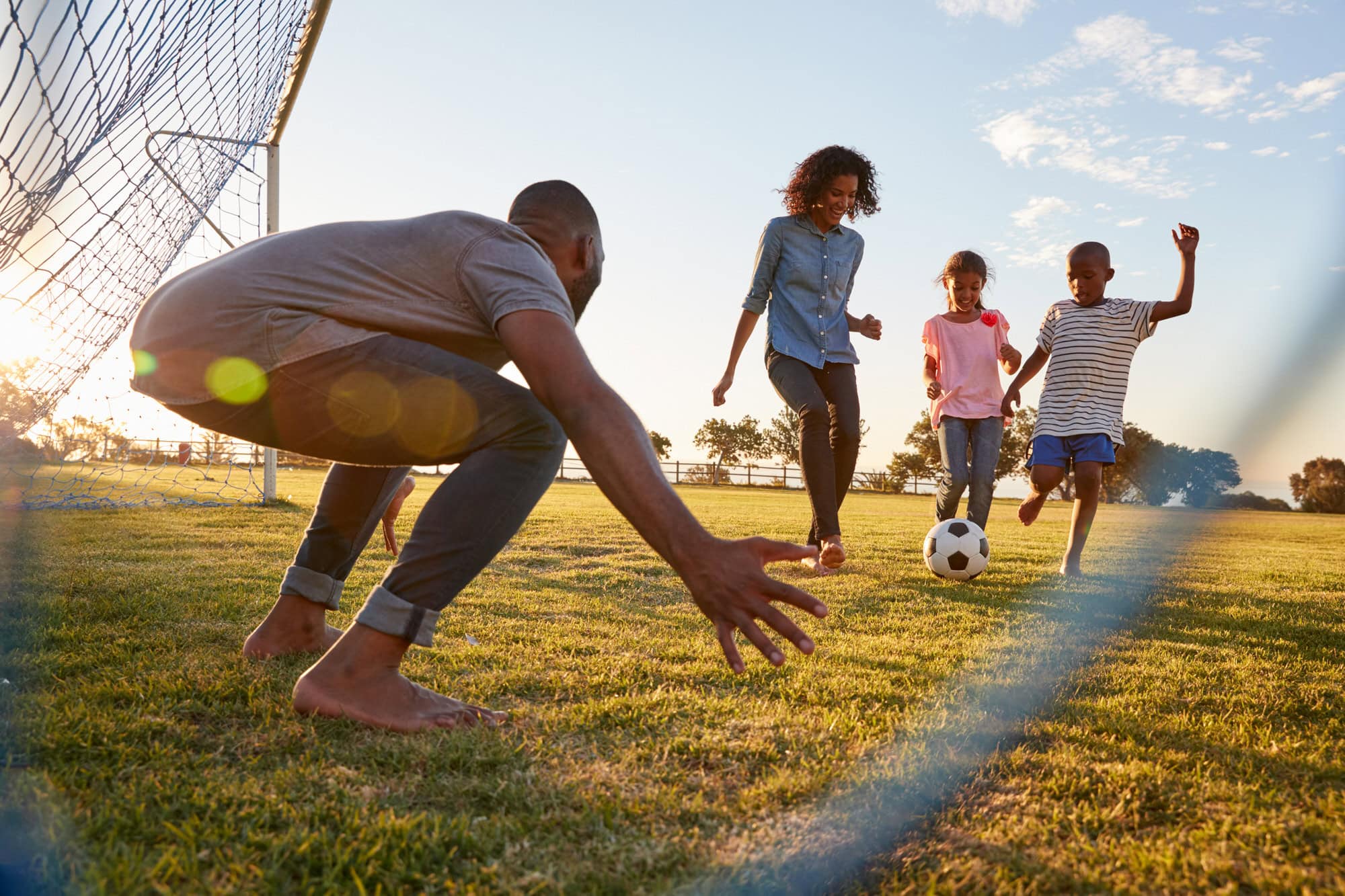 A family of four plays soccer on a grassy field, with an adult acting as goalie and two children chasing the ball while another adult observes.