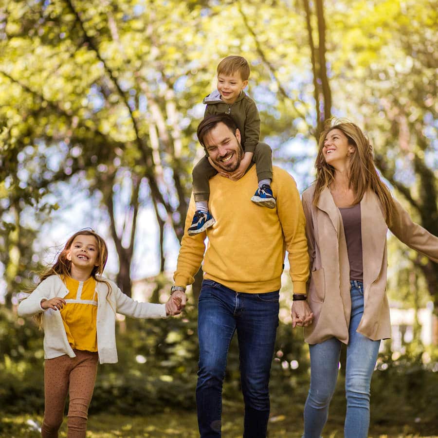 A family of four walks outdoors in a sunlit park; the man carries a young boy on his shoulders, while the woman and a girl walk beside them, all smiling.