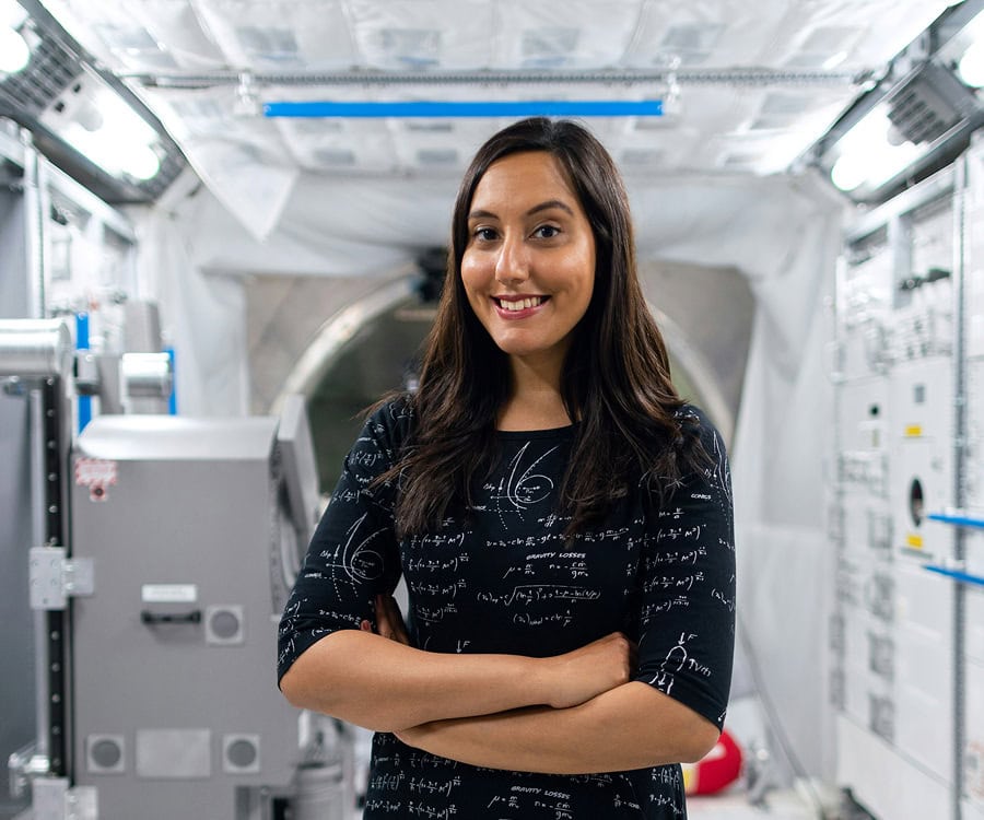 A woman with long dark hair stands confidently with arms crossed in a high-tech, white laboratory or spacecraft interior.