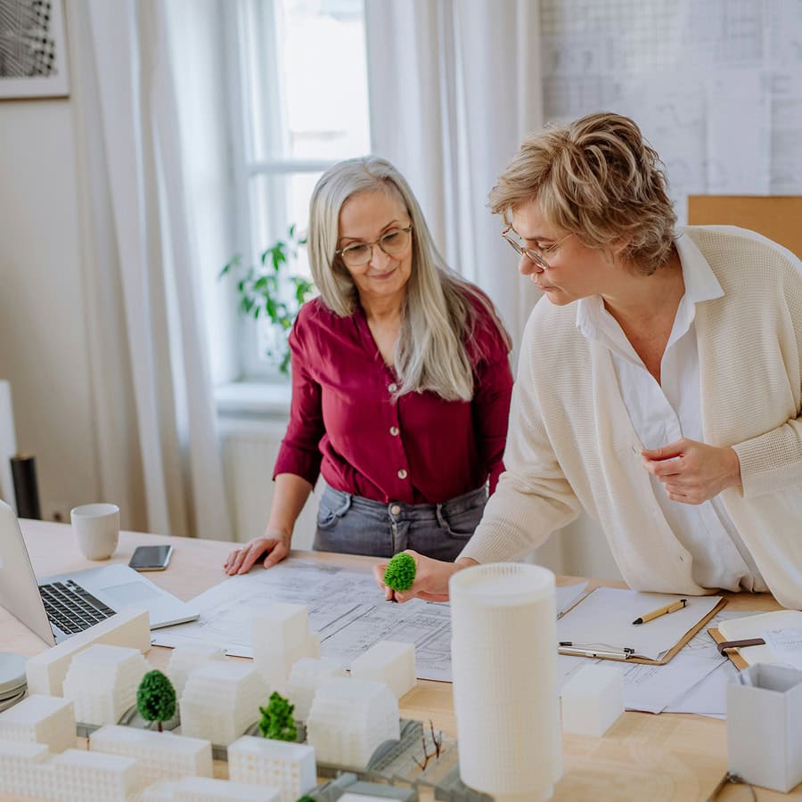 Two women review architectural plans and building models on a table in a bright office, discussing a project with a laptop and paperwork nearby.