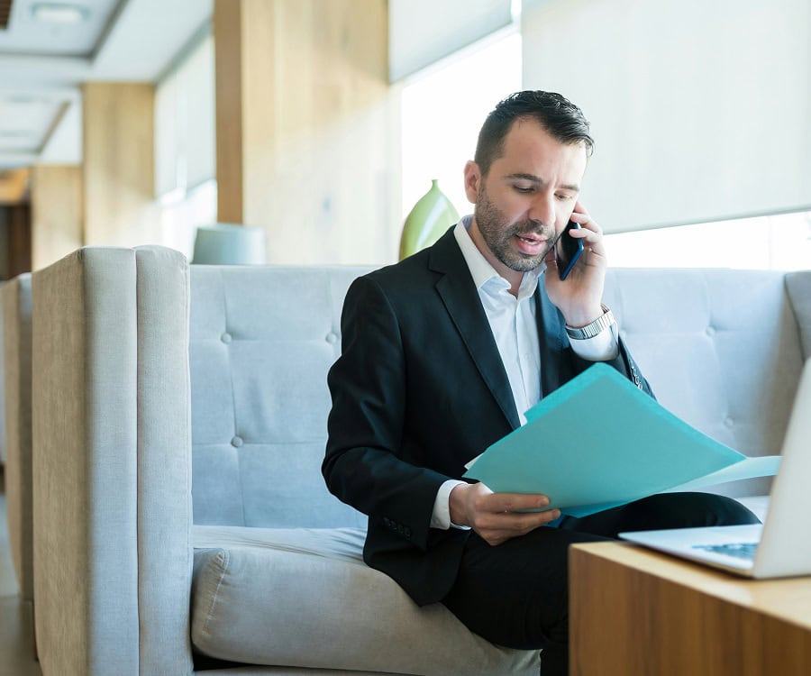 A man in a suit sits on a couch, talking on his phone while reviewing documents and using a laptop on a nearby table.