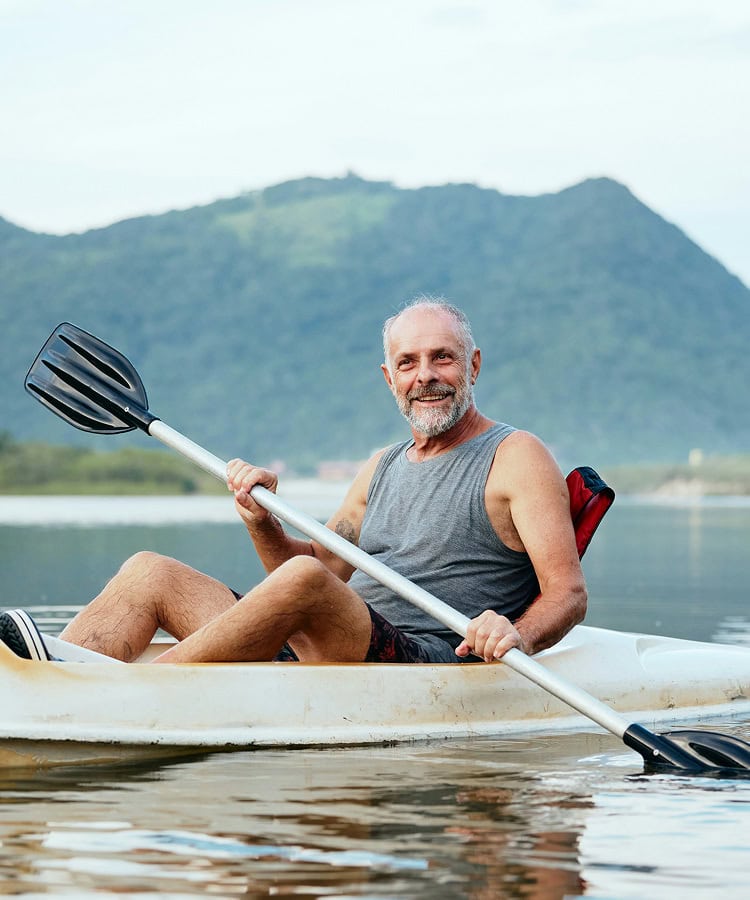 An older man in a sleeveless shirt smiles while kayaking on calm water, with green hills and a cloudy sky in the background.
