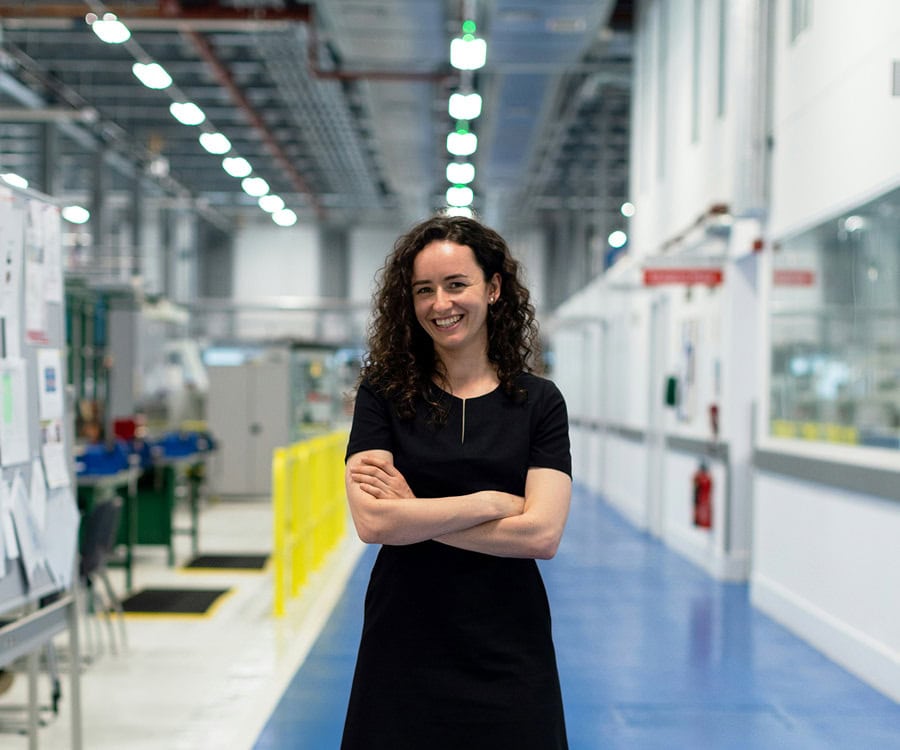 A woman with curly hair stands with arms crossed and smiles in a brightly lit industrial facility or laboratory hallway.