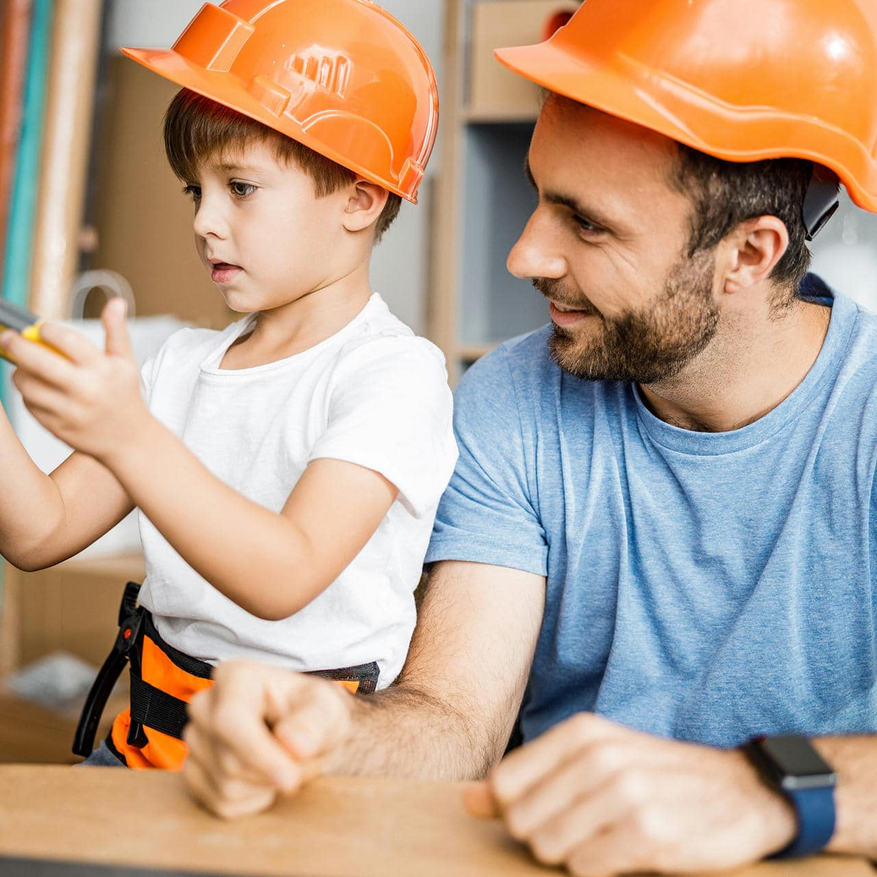 An adult and a child wearing orange construction helmets sit together. The child holds a tool and is focused on it, while the adult looks on and smiles.