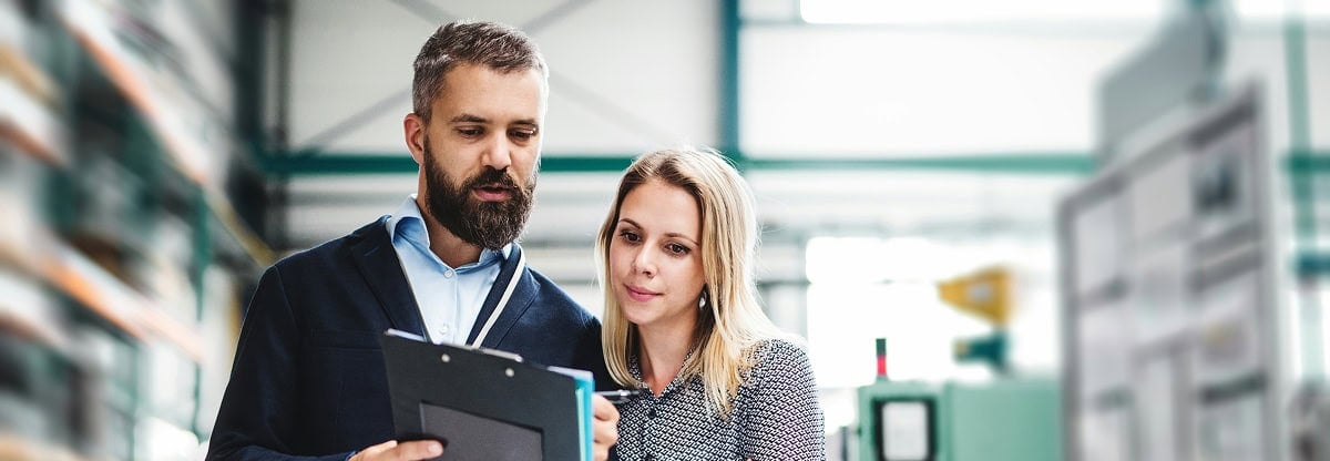 Two people standing indoors review documents on a clipboard, surrounded by industrial shelving and equipment in a warehouse or factory setting.
