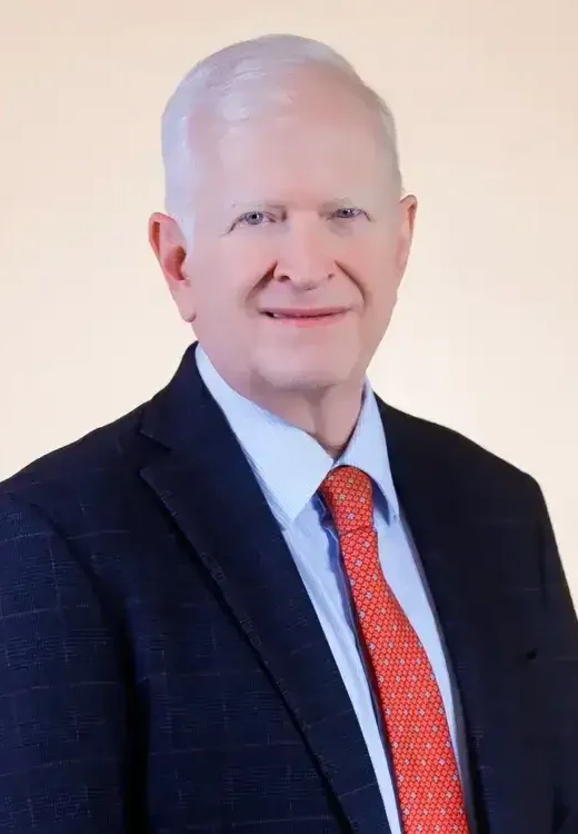 An older man with short white hair wearing a dark suit, light blue shirt, and red tie, standing in front of a plain light-colored background.