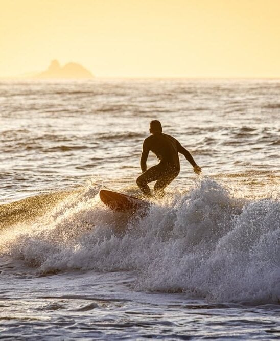 A surfer rides a wave on the ocean at sunset, with distant rocks and a boat visible on the horizon.