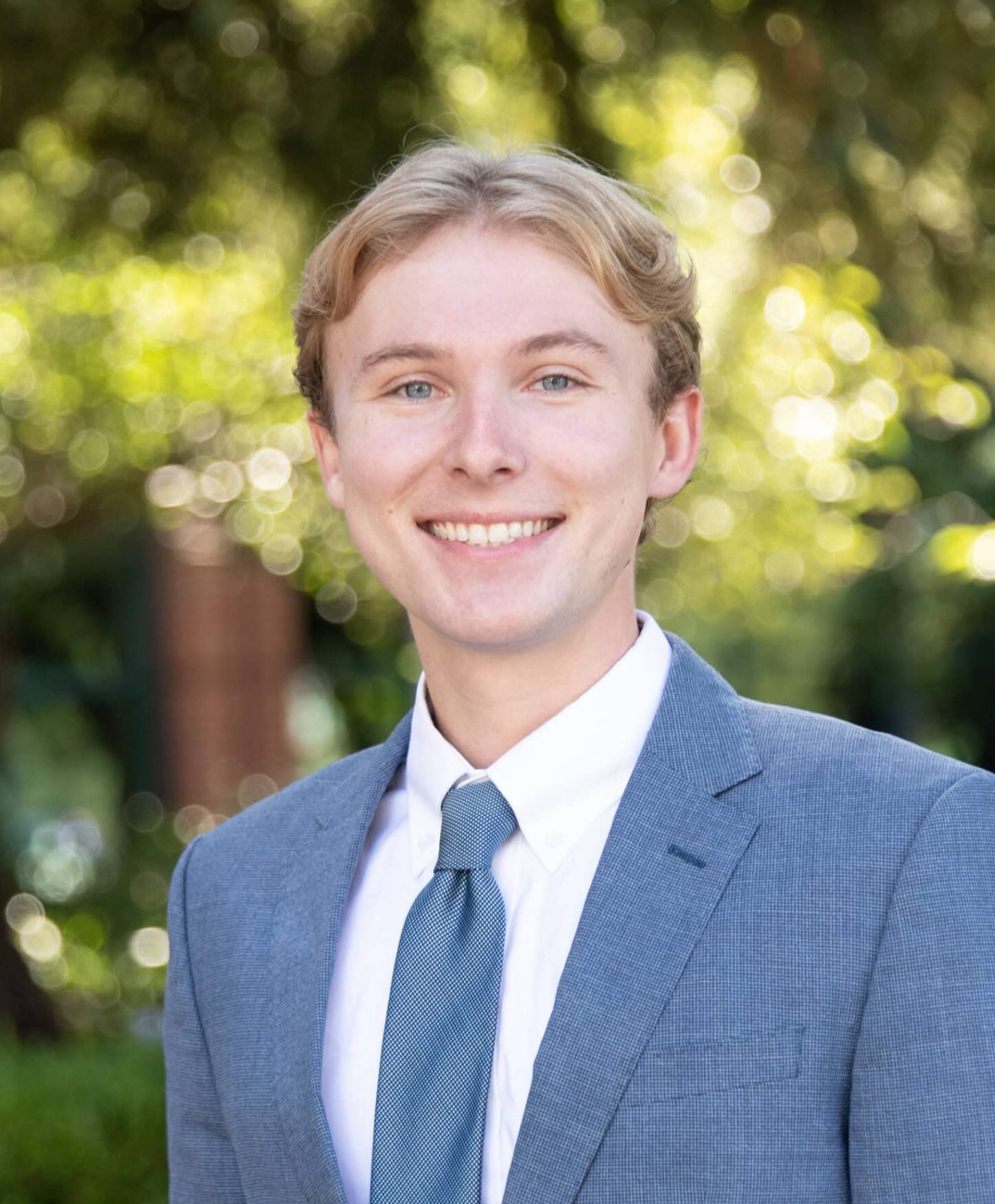 Young man in a light gray suit and blue tie smiles at the camera outdoors, surrounded by greenery and sunlight in the background.